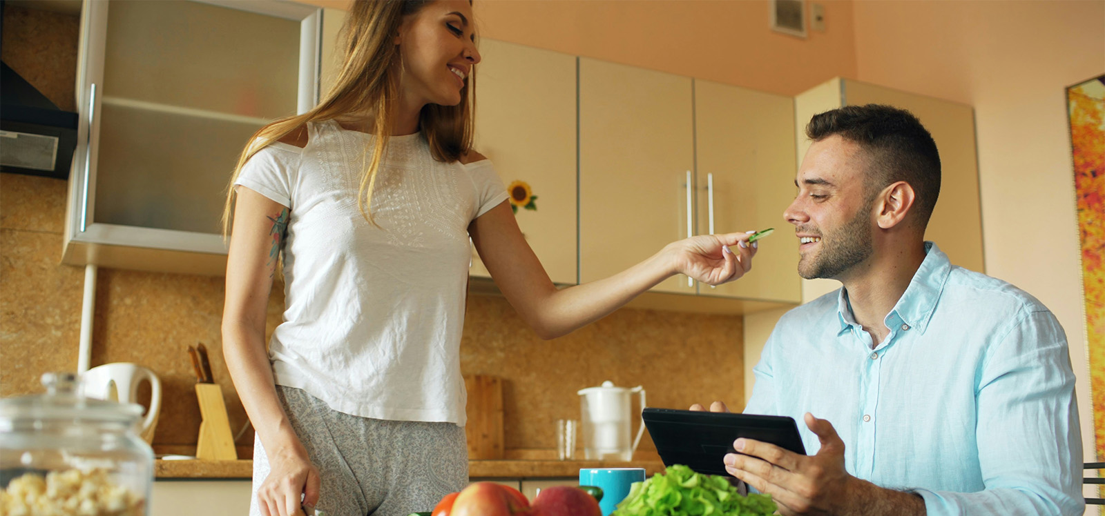 A blonde woman offering her spouse a cucumber slice